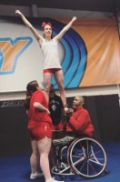 Adaptive abilities athletes performing a stunt. One base is holding a top person at her waist while the other base is in a wheelchair, holding the top person at his chest.