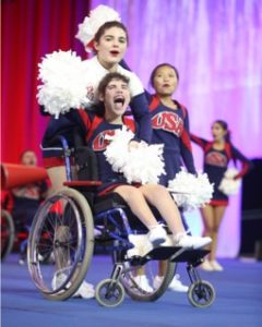 A USA Cheer national team Special Abilities athlete in a wheelchair is holding pom poms and cheering alongside her ambassador at the world championship. 