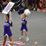 County youth cheering at a competition with signs and poms. They are dressed in purple uniforms with white sleeves.