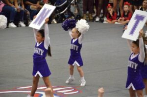 County youth cheering at a competition with signs and poms. They are dressed in purple uniforms with white sleeves.