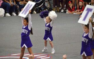 County youth cheering at a competition with signs and poms. They are dressed in purple uniforms with white sleeves.